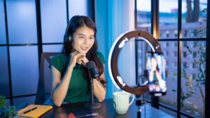 Uma criadora de conteúdo sorridente usando fones de ouvido e falando em frente a um smartphone montado em um ring light (luz circular), em um ambiente de estúdio caseiro.