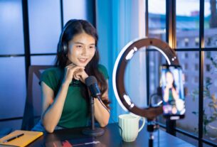 Uma criadora de conteúdo sorridente usando fones de ouvido e falando em frente a um smartphone montado em um ring light (luz circular), em um ambiente de estúdio caseiro.