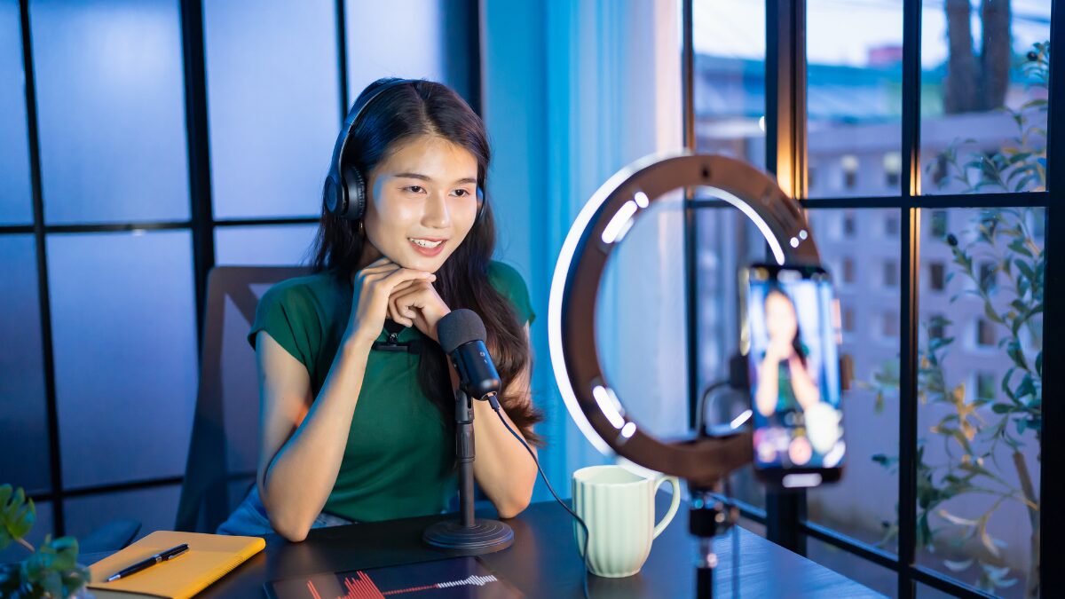 Uma criadora de conteúdo sorridente usando fones de ouvido e falando em frente a um smartphone montado em um ring light (luz circular), em um ambiente de estúdio caseiro.