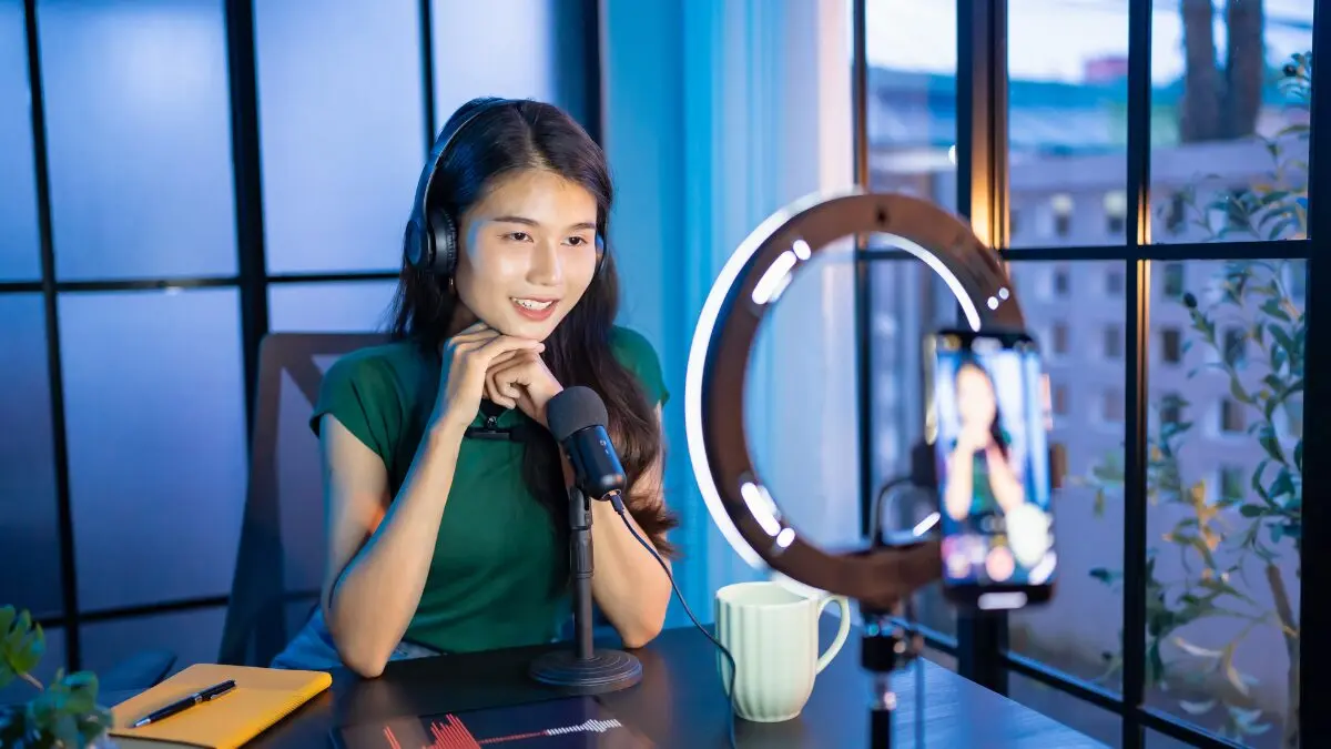 Uma criadora de conteúdo sorridente usando fones de ouvido e falando em frente a um smartphone montado em um ring light (luz circular), em um ambiente de estúdio caseiro.