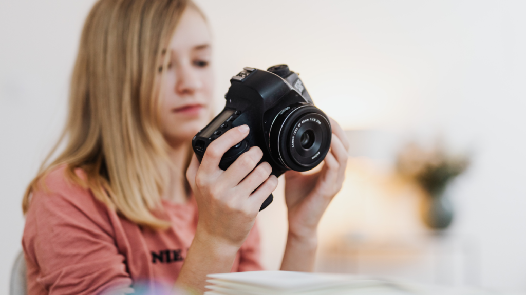 Mulher loira sorridente segurando uma câmera fotográfica DSLR profissional em um ambiente iluminado.