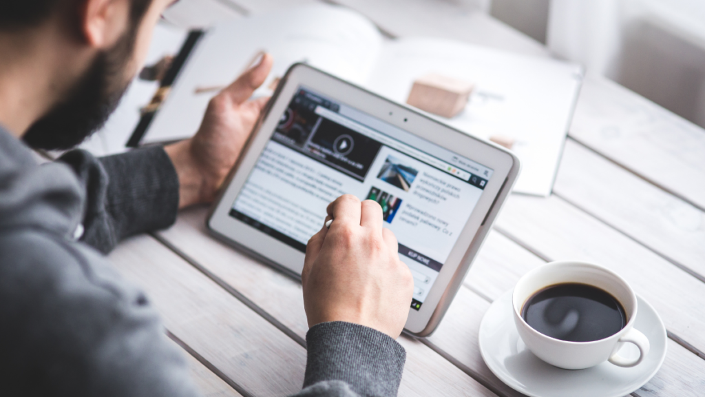 Pessoa utilizando um tablet com gráficos e textos sobre marketing digital, acompanhada de uma xícara de café e caderno.