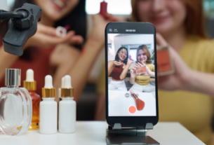 Duas mulheres produzindo conteúdo de marketing digital com smartphone e produtos de beleza sobre a mesa.