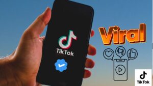 Foto de uma mão segurando um celular com o logotipo do TikTok e um selo azul de conta verificada, ao lado da palavra "Viral" escrita em letras grandes com ícones de engajamento.