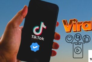 Foto de uma mão segurando um celular com o logotipo do TikTok e um selo azul de conta verificada, ao lado da palavra "Viral" escrita em letras grandes com ícones de engajamento.